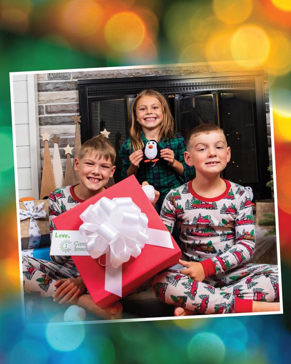 Three siblings wearing festive pajamas sit cross-legged on the floor and hold up a large red gift box and a small penguin stuffie